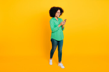 Fototapeta na wymiar Full length photo of positive stylish beautiful brazilian or african american curly haired woman, with glasses, holding her smartphone, typing message, smiling, stand on isolated yellow background