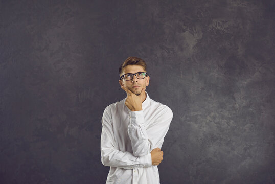 Portrait of serious confused intrigued undecided young man in glasses standing in studio, looking up, holding hand on chin, thinking, doubting, trying to make difficult choice or answer hard question