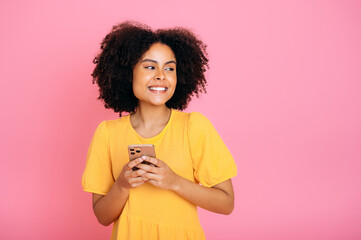 Positive hispanic or brazilian woman, in yellow sundress, using her smartphone, chatting online with friends, browsing internet, texting, looks to the side, smiling, stand on isolated pink background