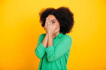Scary, afraid. Terrified hispanic or brazilian curly haired woman dressed in casual shirt, peeks through fingers, covers face with hands, experiencing scare, shock, stand on isolated yellow background