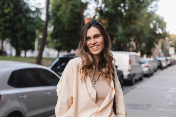 Fototapeta premium Friendly attractive young blonde woman relaxing leaning against a street, cars exterior space in an urban street staring at camera. Happy smiling woman wear yellow shirt, beige to and brown bag.