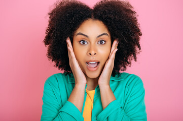 Fototapeta na wymiar Amazed emotional african american or brazilian curly haired beautiful woman in green shirt, looking at camera in surprise, holding palms near cheeks and opening her mouth, isolated pink background