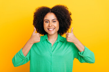 Joyful excited curly haired brazilian or african american stylish positive woman, in green shirt, looks happily at the camera and smile, shows thumb up gesture, stands on isolated yellow background