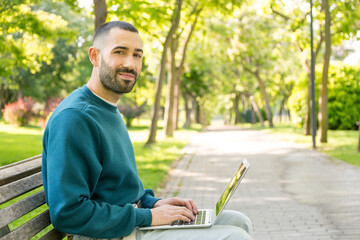 Fototapeta premium Young bearded man smiling, sitting on a park bench with a laptop