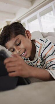 Vertical video of biracial boy lying on sofa and using tablet at home, slow motion