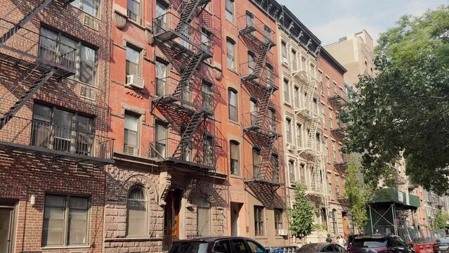 A daytime summer exterior establishing shot of typical red brick apartment buildings with fire escapes in Manhattan or Brooklyn.  	