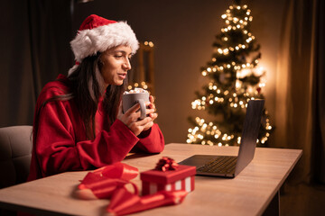 Obraz na płótnie Canvas Young happy woman having video call over laptop while celebrating Christmas alone at home. Watch movie with marshmallow