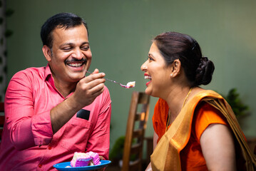 Happy Indian husband feeding cake to wife while sitting on sofa at home - concept of wedding anniversary celebrating, family bonding and affection.