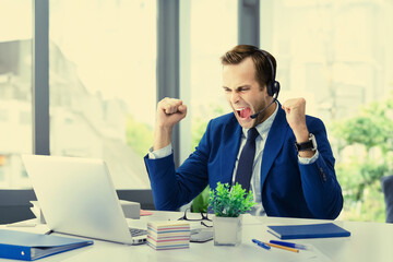 Photo of very happy businessman in headset suit with laptop computer at office. Excited business man raising hands up clenching fists - success concept. Trade mentor coach sitting on window background