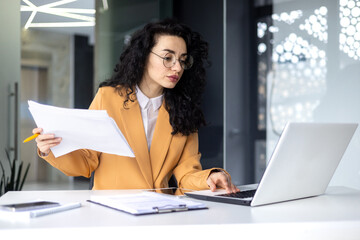 Serious focused female financier working inside office with documents and papers at workplace,...