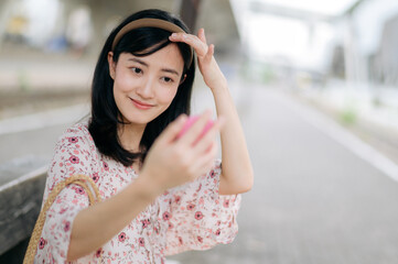 young asian woman traveler checking her face in a mirror and waiting for train in train station. Journey trip lifestyle, world travel explorer or Asia summer tourism concept.