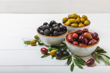 Green, black and red olives on a white wooden background. Various types of olives in bowls and olive oil with fresh olive leaves. Copy space. Place for text. Mediterranean food. Vegan.