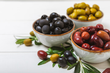 Green, black and red olives on a white wooden background. Various types of olives in bowls and olive oil with fresh olive leaves. Copy space. Place for text. Mediterranean food. Vegan.