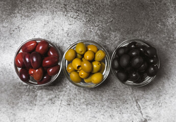 Green, black and red olives on a textured marble background. Various types of olives in bowls and olive oil with fresh olive leaves. Copy space. Place for text. Mediterranean food. Vegan.