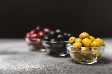 Green, black and red olives on a textured marble background. Various types of olives in bowls and olive oil with fresh olive leaves. Copy space. Place for text. Mediterranean food. Vegan.