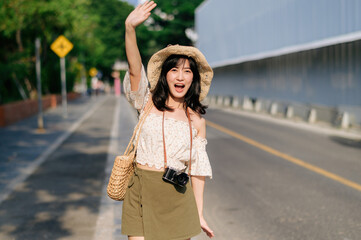 Portrait of asian young woman traveler with weaving hat and basket and a camera waving hand to friend by the street. Journey trip lifestyle, world travel explorer or Asia summer tourism concept.