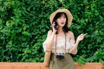 Obraz premium Portrait of asian young woman traveler with weaving hat, basket, mobile phone and camera on green public park background. Journey trip lifestyle, world travel explorer or Asia summer tourism concept.