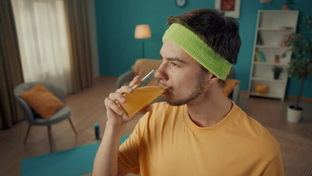 Portrait of a young man with a headband after a home workout. A man is happy to drink freshly squeezed juice, fresh against the backdrop of a living room with attributes for home fitness. Slow motion.