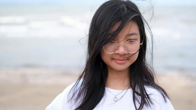 Asian girl wearing eyeglasses in a white t-shirt stands with her hair blowing and smiling cheerfully by the sea.