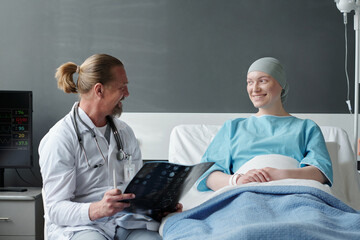 Happy young woman with headscarf sitting in bed and talking to her doctor with brain MRI scan...