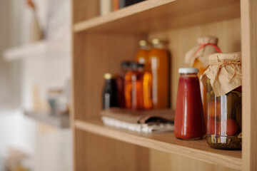 Focus on group of jars with homemade ketchup, pickled tomatoes and other food products standing on wooden shelf in the kitchen