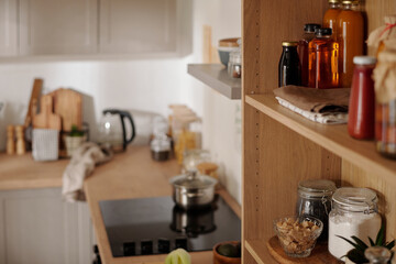 Fototapeta na wymiar Wooden shelves with jars containing flour and coffee beans, bottles with pickles, vegetable oil, homemade juice and ketchup in the kitchen