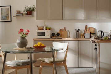 Part of spacious kitchen with glass round table and two wooden chairs standing in the center, grey cabinets on wall and on the floor