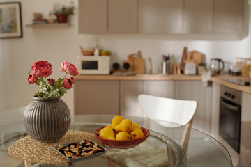 Several yellow pears on plate, nuts and vase with bunch of red flowers on glass round table standing in the center of spacious kitchen