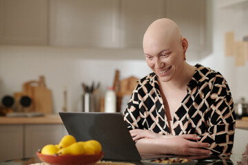 Young smiling bald woman looking at her friend on laptop screen while sitting in the kitchen at home and communicating in video chat
