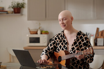 Young smiling woman with acoustic guitar looking at laptop screen while sitting by table in the...