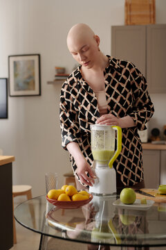 Young bald woman with oncological disease preparing fruit smoothie in electric mixer while standing by kitchen table against paintings on walls