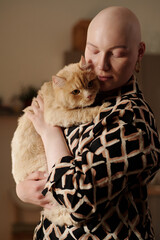 Young affectionate bald female owner of purebred fluffy cat holding her pet on hands while standing...