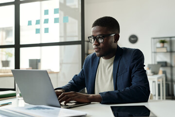 Young serious African American entrepreneur typing on laptop keyboard while sitting by workplace in office and working over new project