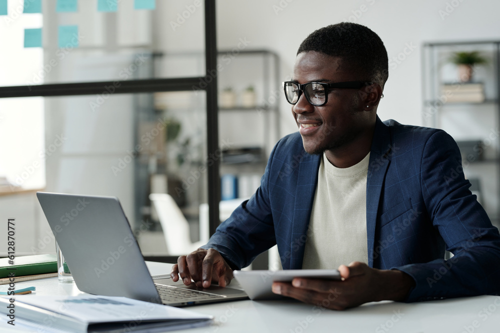 Poster young smiling businessman in eyeglasses looking at laptop screen while analyzing online data or comm