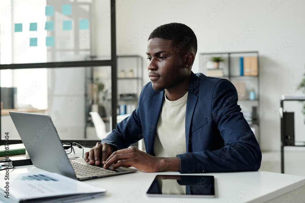 Poster young serious manager in elegant suit looking at laptop screen and analyzing online data or working 