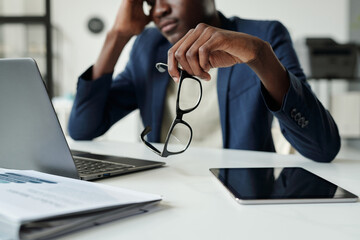 Focus on hand of young pensive African American businessman holding eyeglasses while sitting by desk with laptop and tablet in office