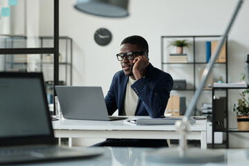 Young confident male broker or agent talking to client on smartphone while sitting by workplace in front of laptop and analyzing data