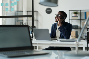 Happy young African American broker talking to client on smartphone while sitting by workplace in front of laptop in openspace office