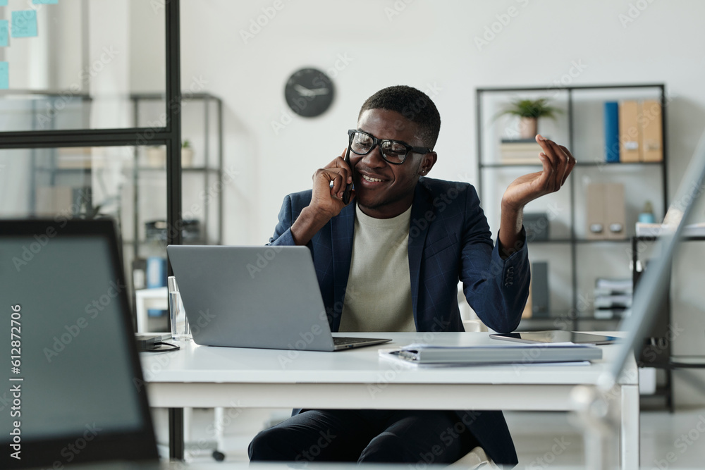 Poster young cheerful broker talking to colleague on mobile phone while sitting by workplace and looking th