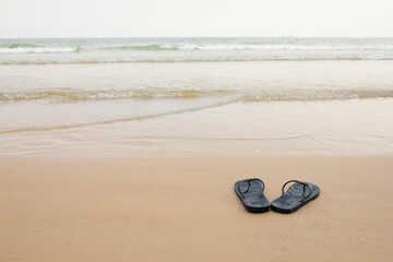 Black slippers feet at the beach, with a wave of foaming gentle beneath them.