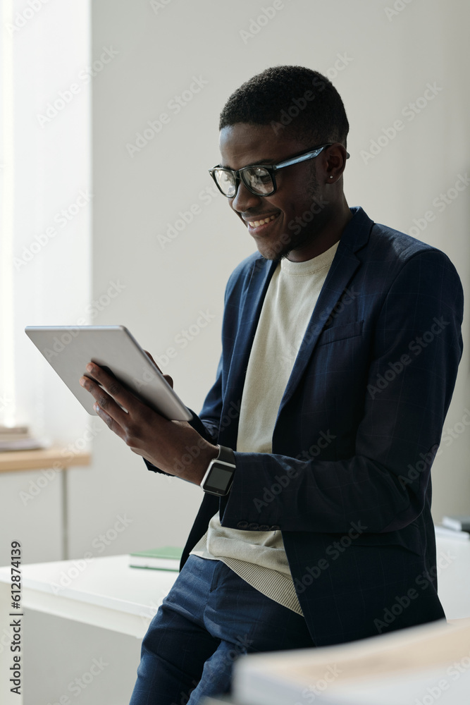 Poster Young smiling male manager in eyeglasses and formalwear using tablet while looking through online information and analyzing it in office