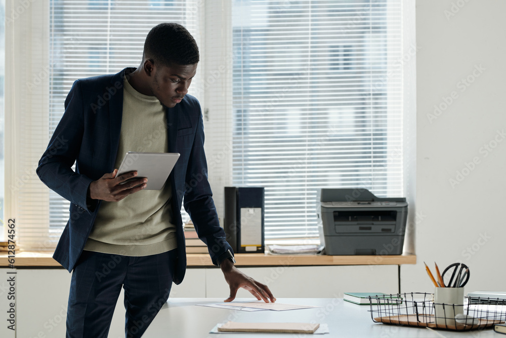 Poster young serious businessman with tablet standing by desk with financial documents in openspace office 
