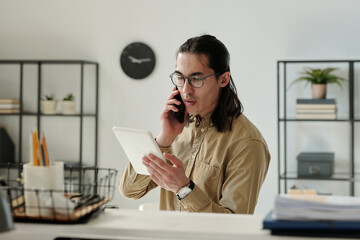 Young serious chief executive officer in eyeglasses and beige shirt talking on smartphone and looking through online data on tablet screen