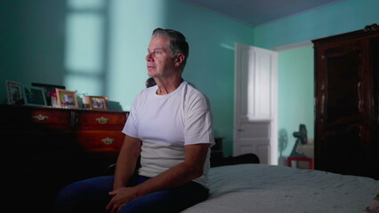 Middle aged man sitting by bed looking at camera in authentic real life domestic lifetyle scene of...