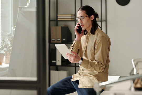 Young chief executive officer in smart casualwear talking to subordinate on mobile phone and explaining online data while looking at tablet screen