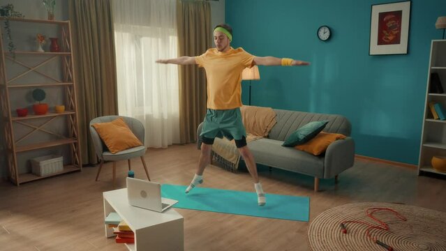 Young man doing star jumps on a gym mat in the living room. A man watches a video lesson on a laptop and repeats the exercises. Home fitness concept, online sports class. Slow motion.