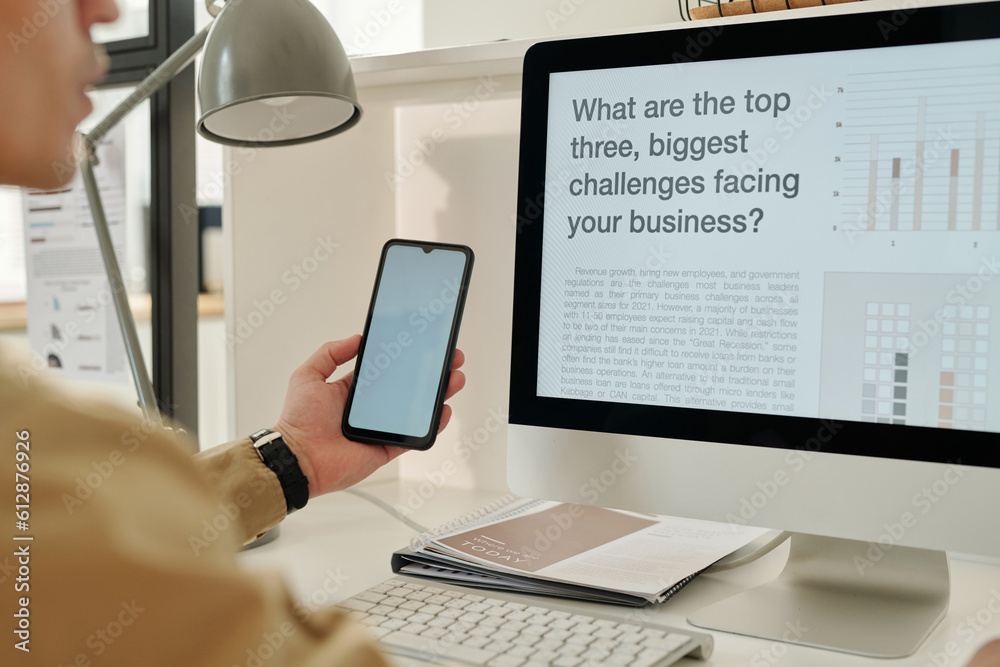 Poster Focus on computer monitor with data on desk and smartphone with blank screen in hand of young male economist sitting by workplace