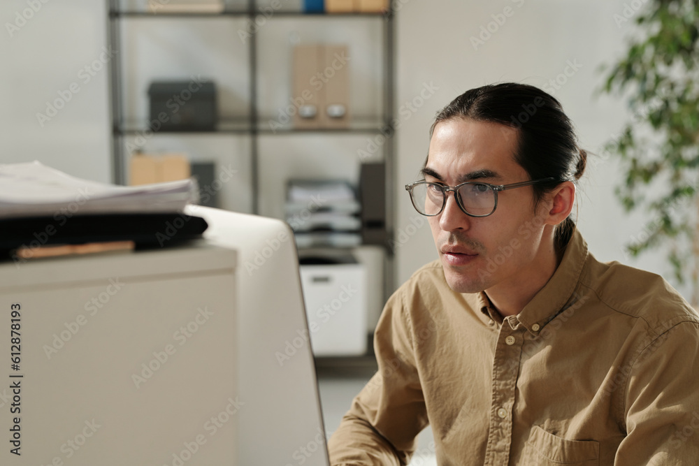 Poster young serious analyst in eyeglasses and beige shirt sitting by workplace in front of computer and lo