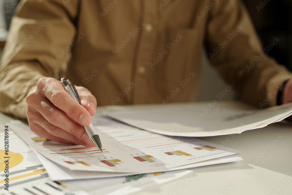 Poster Hand of young economist with pen pointing at financial document with graphic data while analyzing it and preparing report for seminar