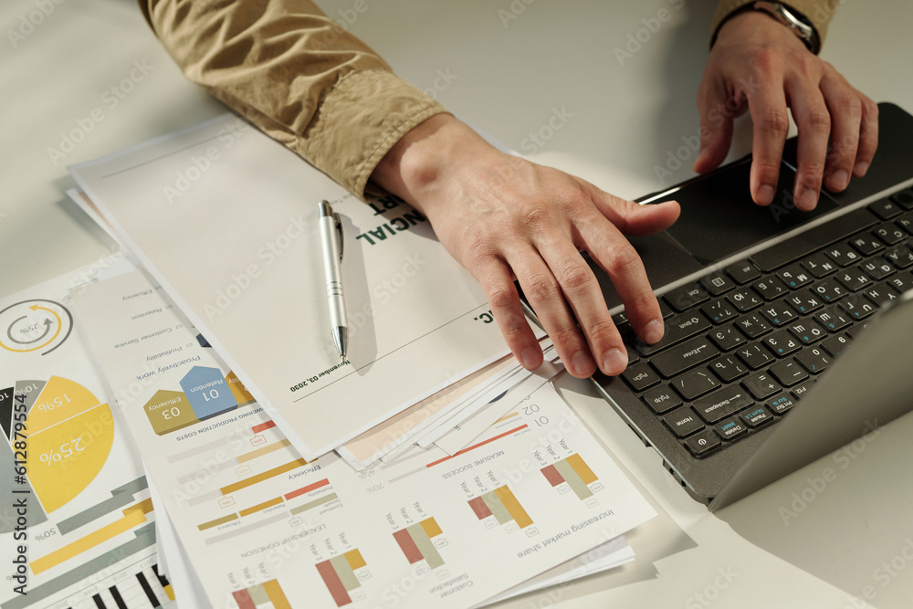 Aufkleber Hands of young male analyst typing on laptop keyboard by workplace while analyzing graphic financial data and entering results in database